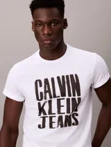Remera blanca de algodón con cuello redondo y manga corta, presentando un estampado gráfico apilado en negro con el texto "CALVIN KLEIN JEANS".