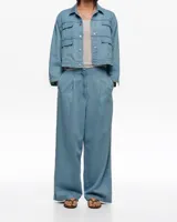 Chaqueta corta estilo boxy de denim azul claro, con cuello camisero, cierre frontal de botones metálicos a presión, mangas largas con puños abotonados y bolsillos con solapa en el pecho.