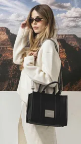 Cartera estructurada de mano color negro, con doble asa superior y correa desmontable. Presenta un diseño minimalista con parche frontal de la marca y costuras verticales decorativas.