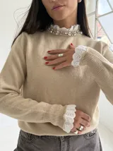 Sweater de punto color beige, con cuello redondo adornado con apliques de pedrería y detalle de puntilla blanca en el cuello y los puños.