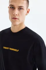 Remera negra de algodón con cuello redondo, mangas cortas y hombros caídos. Presenta un estampado frontal con la frase "TRUST YOURSELF" en color amarillo.
