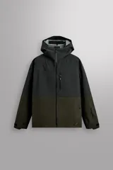 Cazadora deportiva de esquí con diseño bicolor en tonos negro y verde oliva. Presenta capucha amplia con visera, cierre frontal con cremallera termosellada, bolsillos frontales y en manga con cierres impermeables, y aberturas de ventilación en las axilas. Incluye faldón interior antinieve, puños ajustables y tecnología de rescate RECCO.