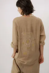Vestido largo de lino color tostado, de manga larga, con escote redondo y diseño amplio y fresco. Cuenta con un delicado bordado en la espalda con motivos marinos y la frase "no se lo digas".