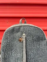 Mochila gris con textura símil cuero, cierre vertical frontal, bolsillo trasero con cierre, interior forrado con bolsillo con cierre y correas regulables.