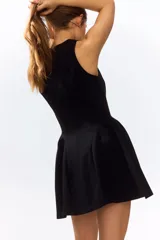 Vestido corto negro sin mangas, con escote redondo, cuerpo entallado y falda acampanada con frunces caídos en la cadera.