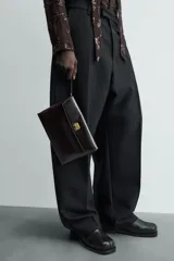 Bolso de mano tipo sobre color marrón oscuro, confeccionado en cuero. Cuenta con cierre de solapa con broche metálico dorado y asa de mano.