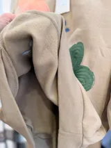 Buzo de cuello redondo en color beige, con diseño oversize y felpa interna. Presenta bordados de flores en tonos rosa, verde y azul distribuidos en el frente.