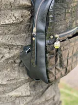 Mochila de cuero sintético color negro con diseño combinado: parte superior con textura de reptil y parte inferior con diseño acolchado capitoneado. Cuenta con bolsillo frontal con cierre metálico y tirador decorativo.
