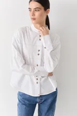 Camisa de corte recto y holgado, color blanco, con cuello mao y cierre frontal de botones. Presenta costuras a la vista en color contrastante, bolsillo lateral tipo parche y puños anchos con detalle bordado.