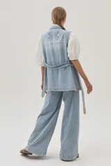 Chaleco largo de jean celeste con cuello camisero, sin mangas, con bolsillos frontales, laterales e internos, y lazo ajustable en la cintura.