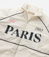 Campera bomber de microfibra color beige con estampado frontal de lettering 'PARIS 1985'. Presenta cierre frontal con cremallera, cuello alto, bolsillos funcionales y puños y ruedo ajustados con elástico.