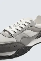 Zapatilla deportiva de hombre con diseño retro, confeccionada en una combinación de materiales que incluye piezas de malla y detalles en cuero con acabado serraje en tonos grises. Presenta cierre de cordones blancos, entresuela blanca en contraste y suela de goma negra con relieve.
