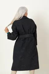 Trench negro capitoneado con bordado floral, corte clásico, cuello solapa, cierre cruzado con broches, lazo para ajustar en la cintura y bolsillos laterales.