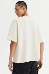 Remera de algodón en punto liviano color beige, con diseño boxy de hombros caídos, cuello redondo con ribete acanalado y corte oversized.