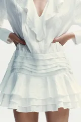 Vestido corto blanco de escote en V y manga larga con puños abotonados. Presenta volantes con bordados de encaje en el cuello, escote y falda. Cierre lateral con cremallera oculta.