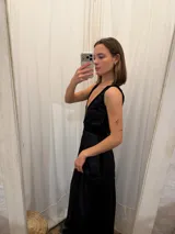 Vestido largo de seda color bronce con acabado brillante, escote profundo en V y espalda abierta. Presenta detalles en terciopelo negro que enmarcan el escote, recorren el frente y definen la cintura con una faja ajustable. Posee breteles anchos y una abertura frontal en la falda.