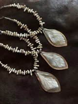 Collar corto con dije central en forma de hoja, elaborado en guampa con engarce de alpaca. La cadena está compuesta por bastoncitos de acrílico y mostacillas transparentes, finalizando con una cadena de eslabones de acero quirúrgico ajustable.
