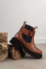 Bota tipo Chelsea de cuero marrón con suela track negra de goma. Presenta paneles elásticos laterales y una costura reforzada en la puntera. Incluye una correa de cuero desmontable con hebilla metálica.