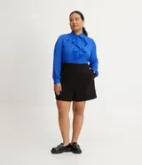 Bermuda negra de crepé, corte amplio y tiro alto, con pliegues frontales y botones dorados decorativos en los bolsillos laterales.