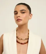 Collar de doble capa con diseño de tubitos curvos. Una de las cadenas presenta tubitos con acabado dorado brillante, mientras que la otra cuenta con tubitos de resina translúcida en tono ámbar, ambos intercalados con pequeñas cuentas metálicas. Posee un cierre de mosquetón y está confeccionado con materiales reciclados.