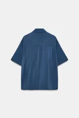 Camisa de corte fluido con cuello solapa y manga corta con dobladillo. Presenta un cierre frontal con botones ocultos bajo una tapeta y un diseño con bajo ligeramente más largo en la parte posterior.