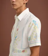 Camisa blanca de lino y algodón, de corte comfort, cuello inglés y abotonado frontal. Presenta manga corta y un bordado floral multicolor con motivos de aves y flores.