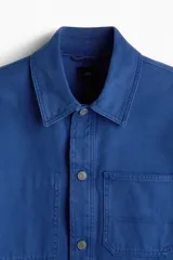 Sobrecamisa de lona de algodón en color azul, con cuello clásico, cierre frontal de botones y tres bolsillos de parche en la parte delantera. Presenta mangas largas con puños abotonados y canesú en la espalda.