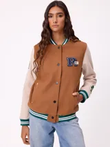 Campera estilo varsity con cuerpo en color marrón y mangas en tono beige. Presenta cierre frontal con botones a presión, cuello, puños y ruedo con franjas tejidas en verde y blanco. Incluye un parche con la letra R en el pecho y detalles bordados en las mangas.