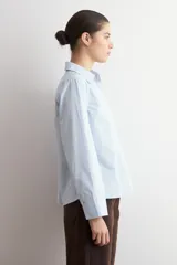 Blusa de algodón color celeste, con cuello camisero, escote en V y mangas largas con puños anchos. Presenta un diseño de corte recto y holgado.