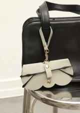 Funda para lentes de cuero sintético color gris claro con detalle de borde festoneado y cierre con broche a presión. Incluye tira para colgar de un bolso.