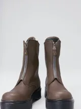 Botas de cuero color marrón de caña alta, con plataforma gruesa de goma y cierre metálico frontal con tirador. Presentan costuras a contratono y detalle de etiqueta de la marca en la lengüeta.