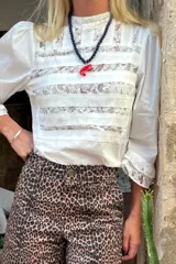 Blusa blanca confeccionada en algodón con inserciones de encaje floral. Presenta un diseño de franjas horizontales en el torso, cuello alto y mangas 3/4 con terminaciones de encaje delicadas.