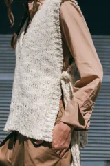 Chaleco tejido de lana rústica en color crudo, con cuello en V y aberturas laterales ajustables con lazos.