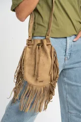 Cartera tipo bucket bag color camel, confeccionada en material sintético con acabado gamuzado. Presenta un diseño decorado con flecos largos en todo el contorno, cierre mediante cordón ajustable y correa larga regulable.
