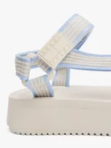 Sandalias de plataforma con suela gruesa dentada color crema. Presentan tiras de tejido trenzado en tonos beige y celeste claro, con cierre adherente y parche tonal de marca en la tira.