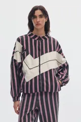 Campera de denim oversize con cierre frontal, cuello alto y bolsillos delanteros. Presenta un diseño a rayas verticales en tonos negros y rosados, cruzado por una franja ancha horizontal de color blanco con forma de zigzag.
