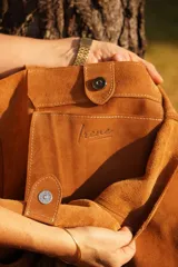 Cartera tipo tote de gamuza color marrón, con diseño amplio y relajado. Presenta un bolsillo frontal externo con cierre de broche metálico y una hebilla decorativa circular, además de un detalle de borla colgante en el lateral.