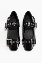Zapatos planos tipo bailarina o mary jane, color negro charol, con punta redonda. Presentan una tira sobre el empeine adornada con múltiples ojales metálicos y una hebilla grande de metal.