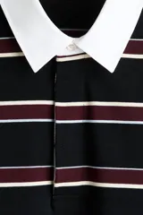 Remera manga larga de punto de algodón con diseño a rayas horizontales en negro, bordó y blanco. Presenta cuello estilo polo en color blanco, cierre frontal parcial con botones ocultos y puños acanalados.