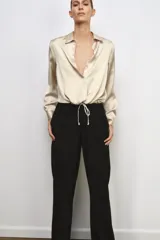 Camisa de cuello solapa y manga larga con puños anchos, confeccionada en tejido satinado color beige. Presenta cierre frontal con botones ocultos y bajo ajustable mediante cordones del mismo tejido.