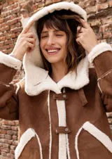 Campera estilo gamulán color tostado con interior y bordes de corderito sintético color crema. Presenta capucha amplia, cierre frontal con trabas y broches metálicos, y bolsillos delanteros grandes. Posee un calce relajado y diseño de textura suave.