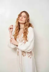 Vestido largo color crudo con bordados florales en tono tostado, escote en V profundo y mangas largas abullonadas con puños elásticos. Incluye cinto a juego.