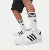 Championes Adidas Superstar de diseño clásico, con capellada de cuero blanco, icónica puntera de goma tipo Shell Toe y las tres bandas laterales en color negro. Presenta detalle en negro en el talón con el logo de la marca.