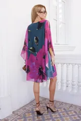 Vestido corto de corte amplio y fluido, con mangas tipo capa y escote redondo. Presenta un estampado floral de grandes flores fucsias sobre un fondo azul oscuro.
