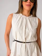 Blusa blanca sin mangas, con cuello trenzado fruncido, detalles de volados en el ruedo y un cinturón fino marrón incorporado.
