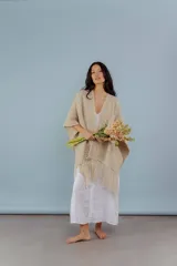 Ruana tejida a mano en lana merino color beige, con textura de punto y detalle de flecos en el ruedo. Presenta sutiles destellos brillantes integrados en el tejido.