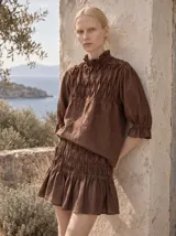 Blusa holgada de lino color marrón, con diseño de nido de abeja fruncido a mano en el canesú delantero, cuello alto con volados, mangas tres cuartos abullonadas con puños elásticos y cierre posterior con botones forrados.