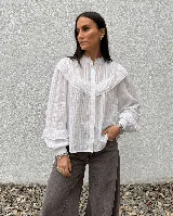 Camisa blanca de algodón con textura a rayas verticales, cuello alto con volados, canesú delantero con volados y encaje, y mangas largas abullonadas con puños ajustados.