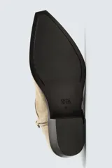 Bota de piel con acabado serraje color beige, con detalle de arnés y hebilla lateral. Presenta horma en punta y suela a contraste con ligero tacón.