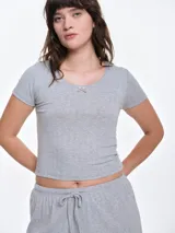 Remera de pijama manga corta en tejido pointelle gris, con escote redondo y detalle de pequeño lazo decorativo en el centro del pecho. Presenta un corte entallado y textura suave.